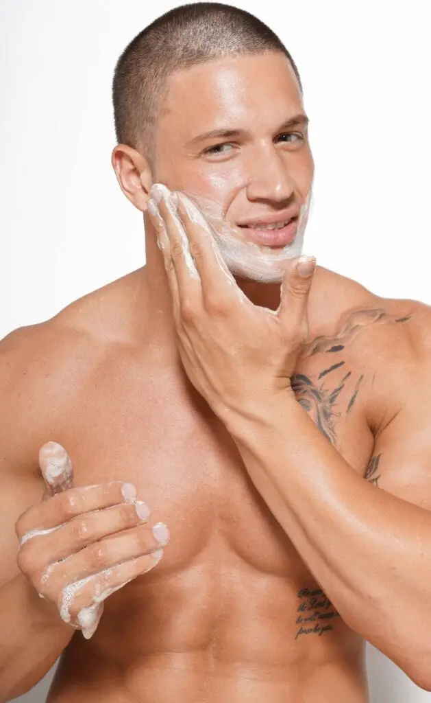 A man with no shirt is shaving his face.